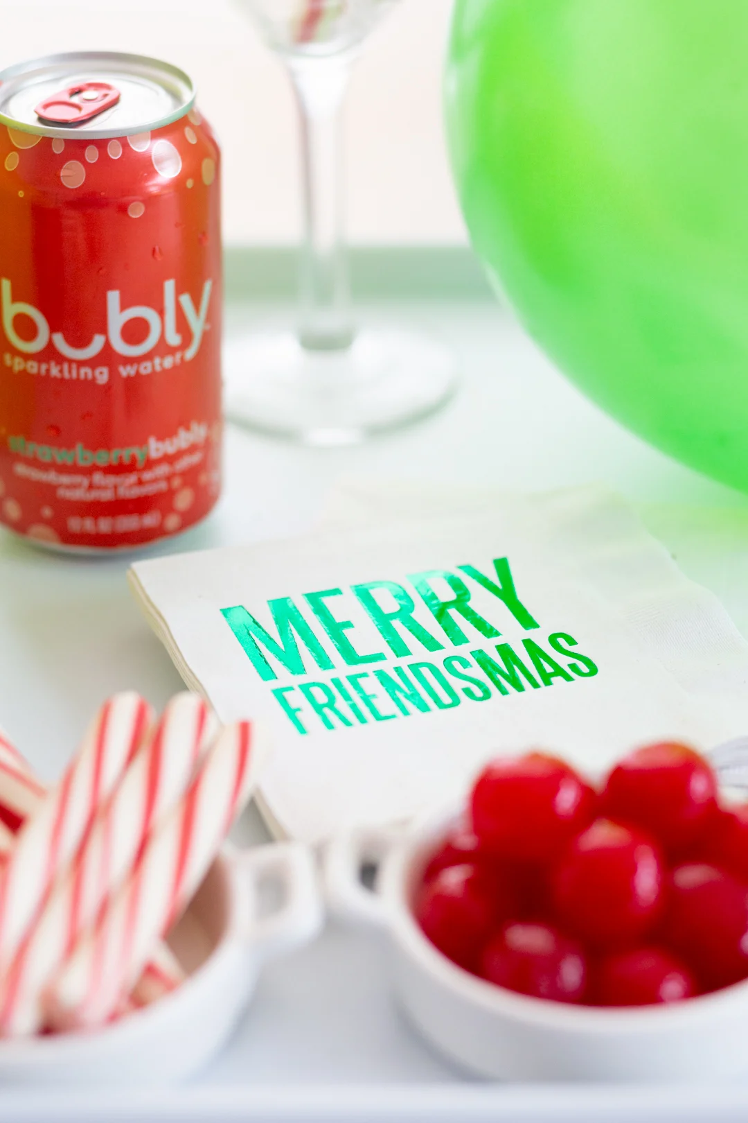 napkins with merry friendsmas written on them surrounded by a drink bar cart items such as a can of sparkling water, peppermint sticks, maraschino cherries and a green balloon.