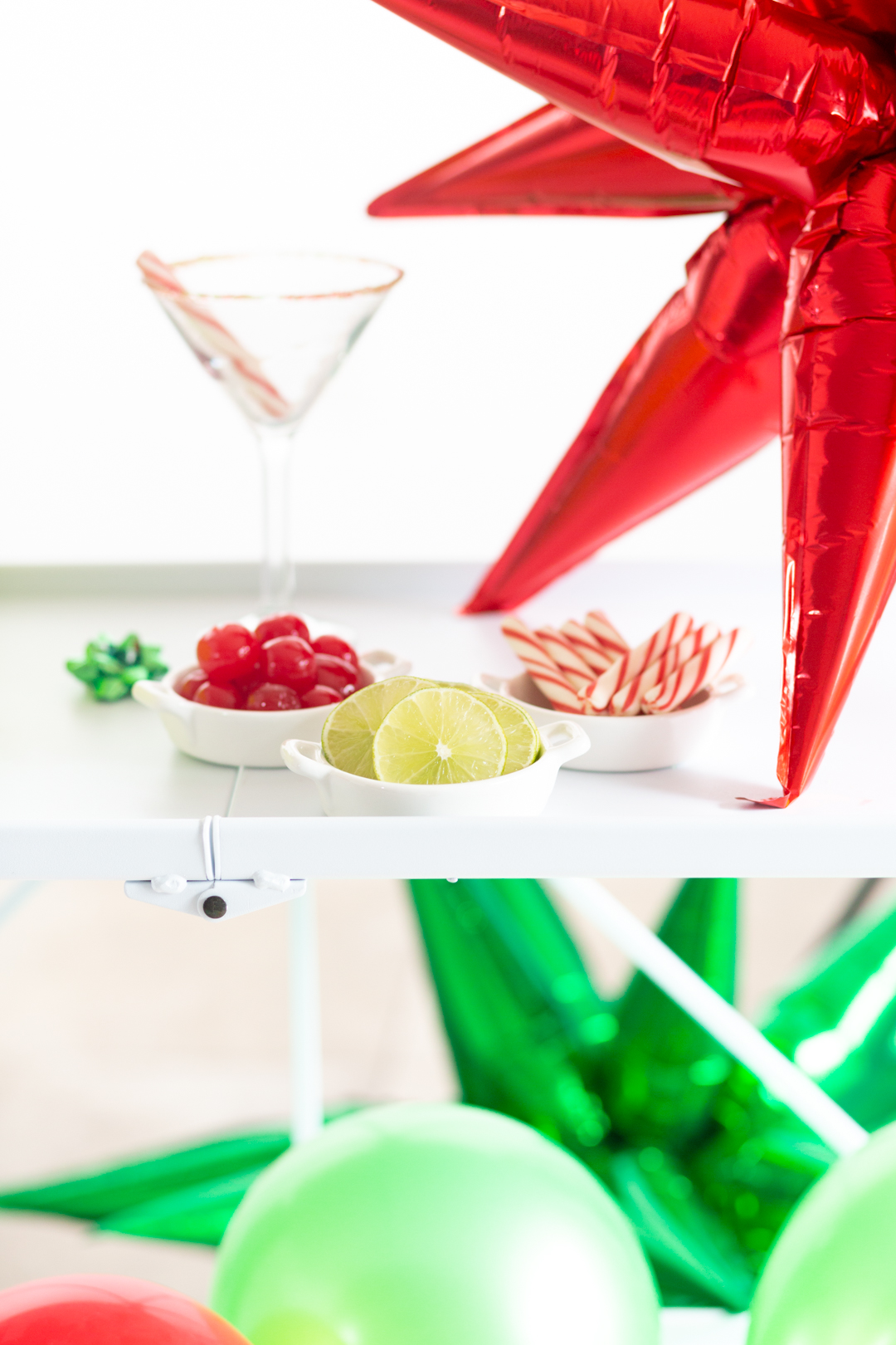 white bar cart with drink garnishes lined up in small white dishes. cocktail glass with sprinkles around the rim in the background. small green bow for decoration on the cart. Large starburst balloons in red and green.