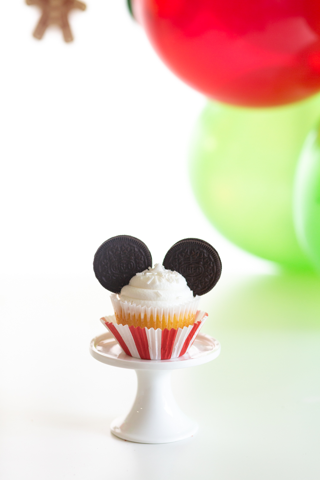 single mickey mouse decorated cupcake for christmas with a red and white striped cupcake liner and two OREO cookies as the ears, white frosting with iridescent sprinkles