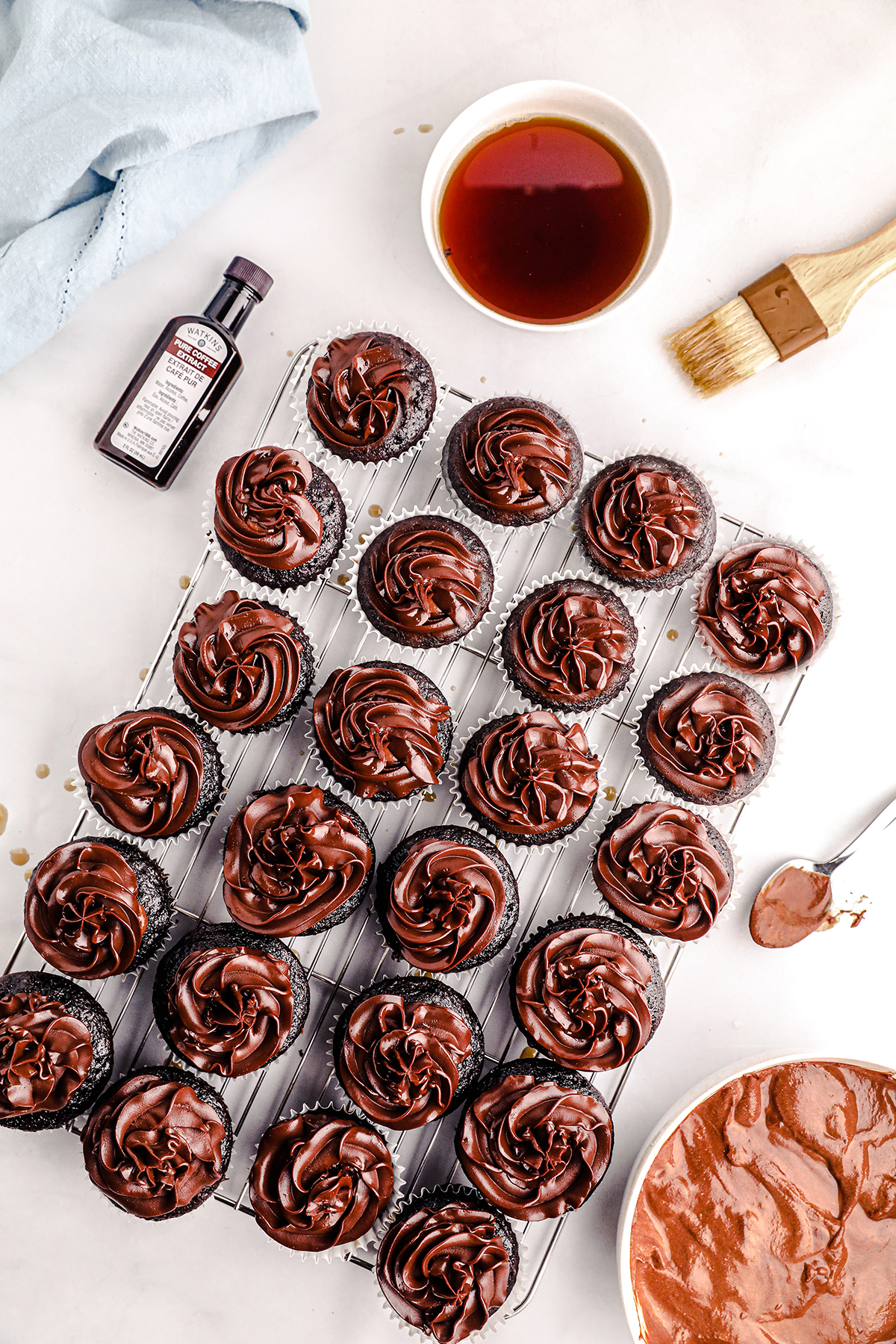 overhead view of batch of chocolate cupcakes with chocolate frosting. vanilla extract nearby and food brush