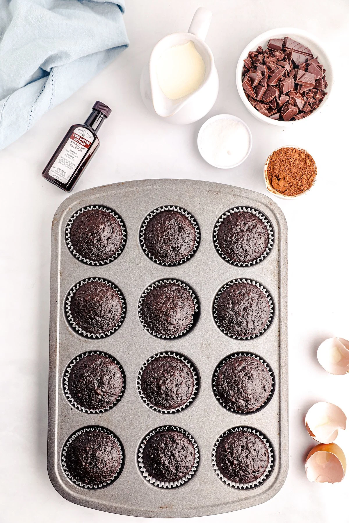 freshly baked chocolate cupcakes out of the oven in a muffin tin. Various ingredients scattered around the tin.