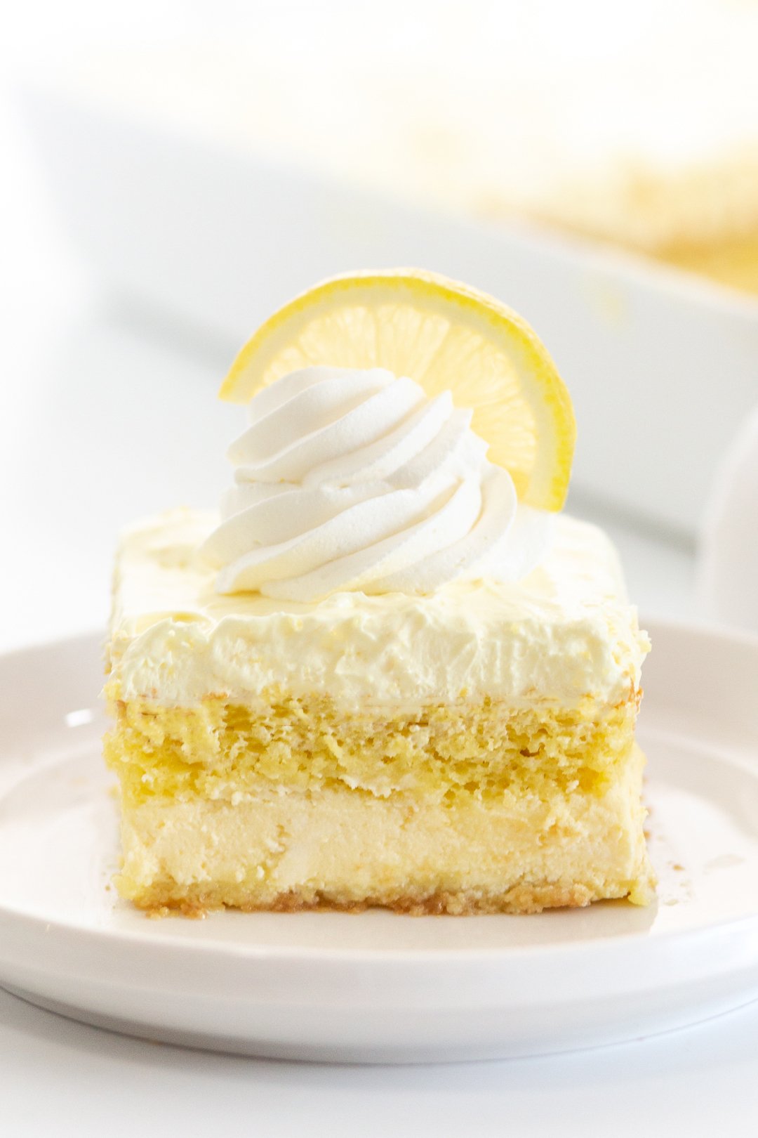 up close view of layered lemon cake on a small white plate. garnished with whipped cream and half slice of lemon