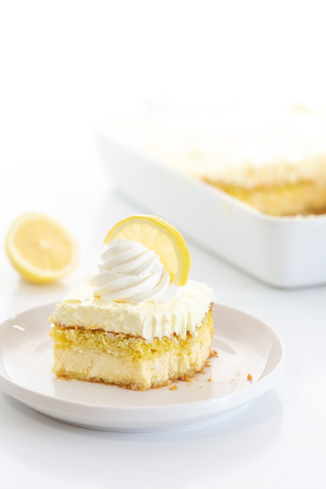 lemon love cake slice served on a small white plate with full cake dish and a halved lemon in the background