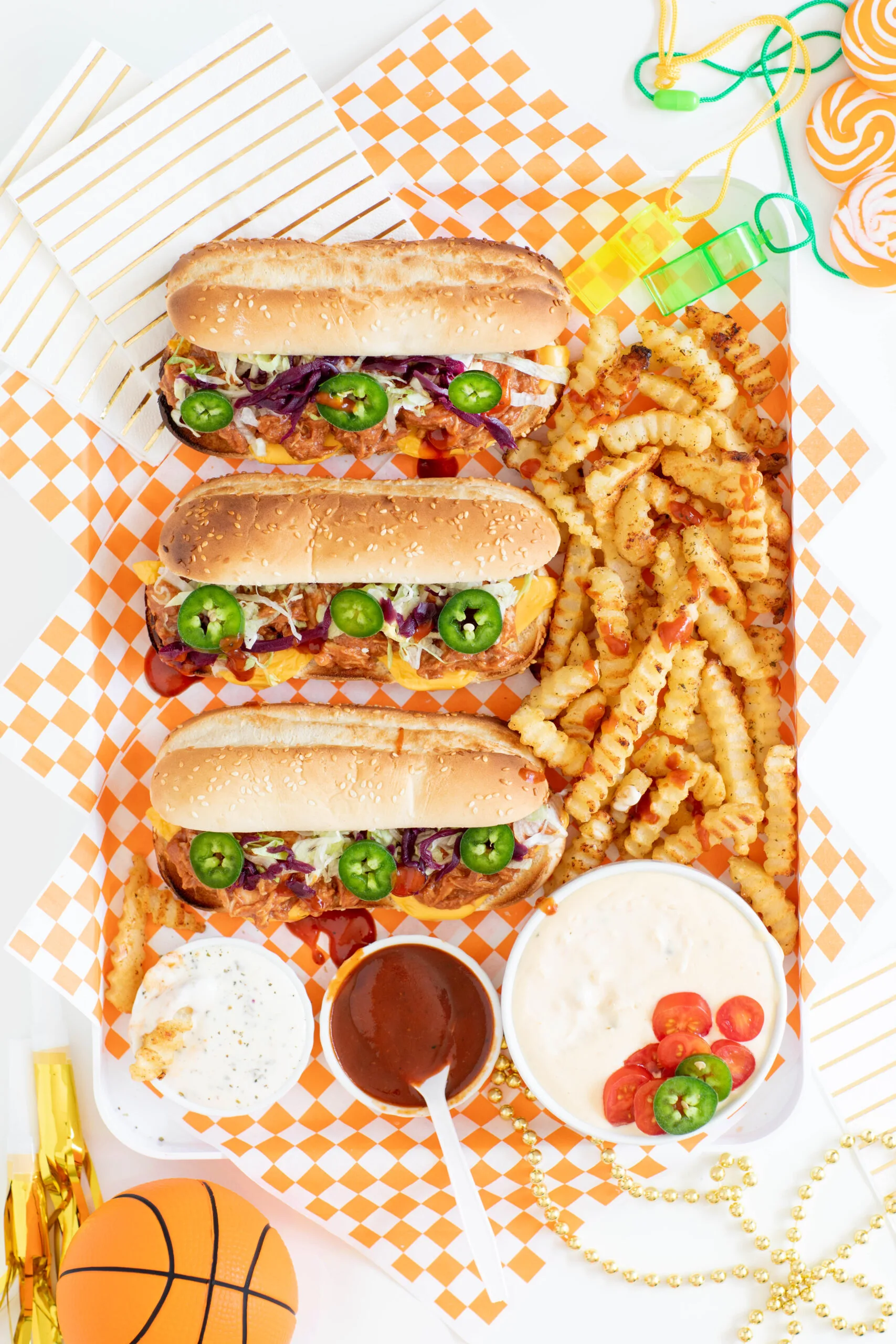 pulled chicken sandwiches layered on a serving tray with crinkle french fries and various condiments and dips in small bowls and dishes. platter is decorated with novelty toy whistles, gold party blowers and mini toy basketball