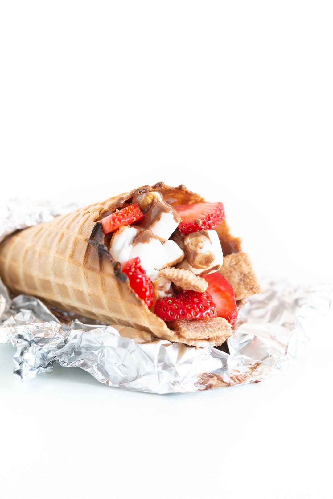 campfire cone resting on foil revealing the melted marshmallows, melted chocolate, strawberry chunks and cinnamon graham cereal inside,