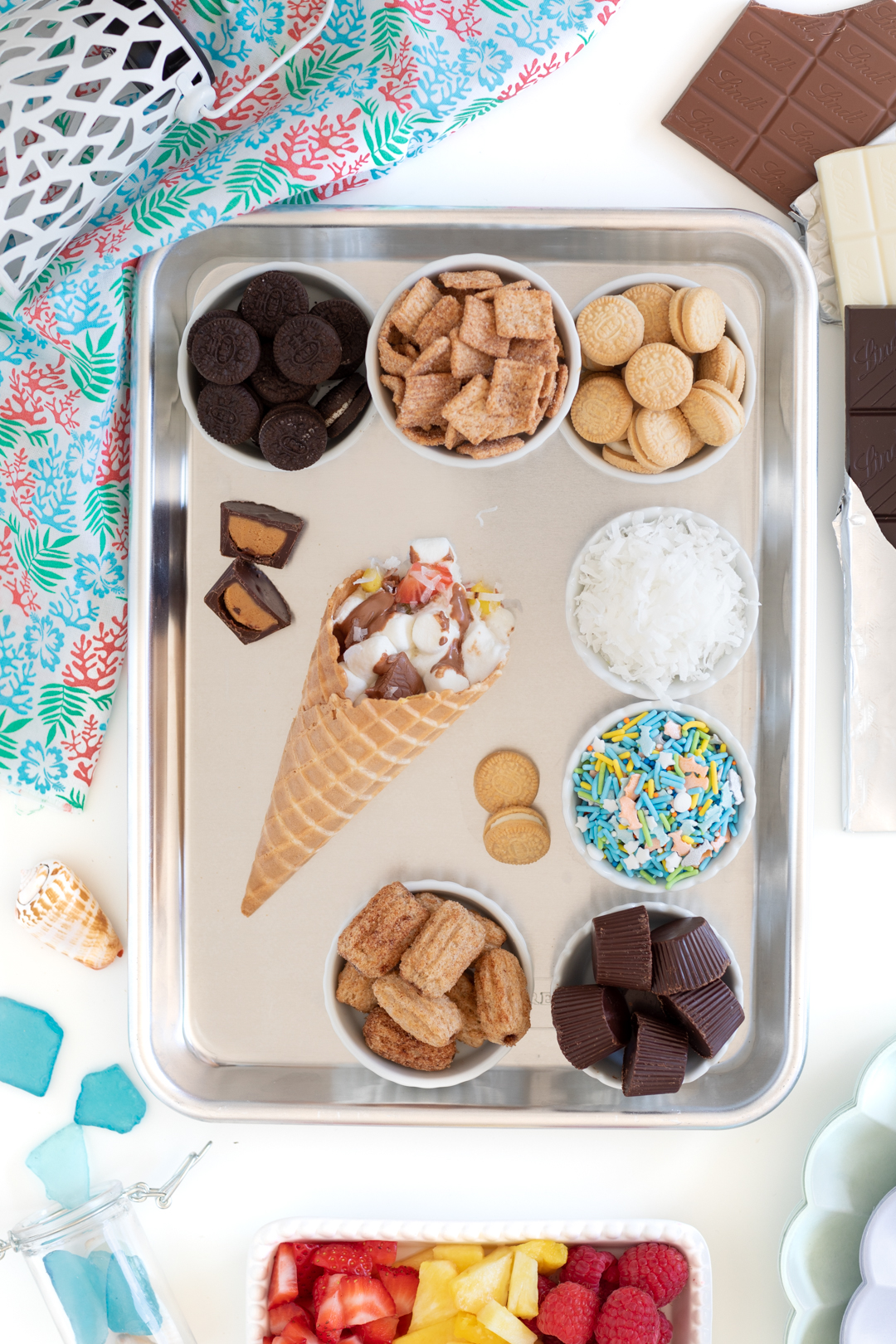 small metal tray filled with campfire cone fixings with ice cream cones and fillings such as mini marshmallows, chocolate, cereals and mini cookies.