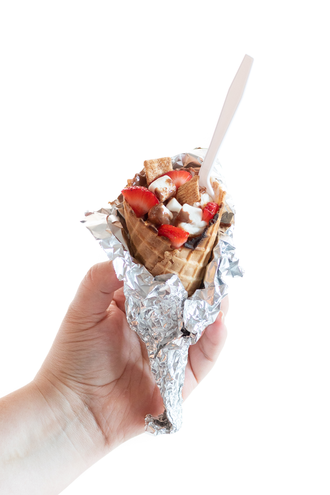 Woman holding a campfire cone wrapped in foil with the top open revealing delicious fillings, chocolate, strawberries, mini marshmallows, cinnamon graham cereal and a spoon on top.