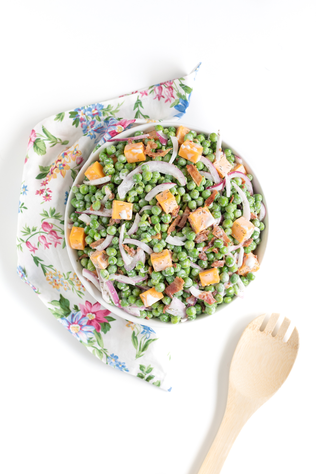 pea salad with bacon, cheese chunks and slivered onions served in a white bowl with floral napkin and serving spoon nearby