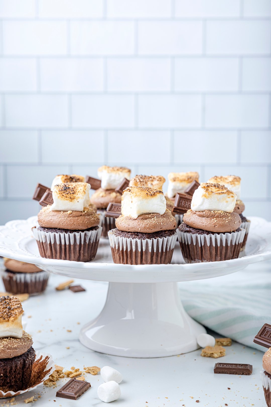 smores cupcakes being served on a white ceramic cake stand. delicious s'mores cupcakes topped with square toasted marshmallows