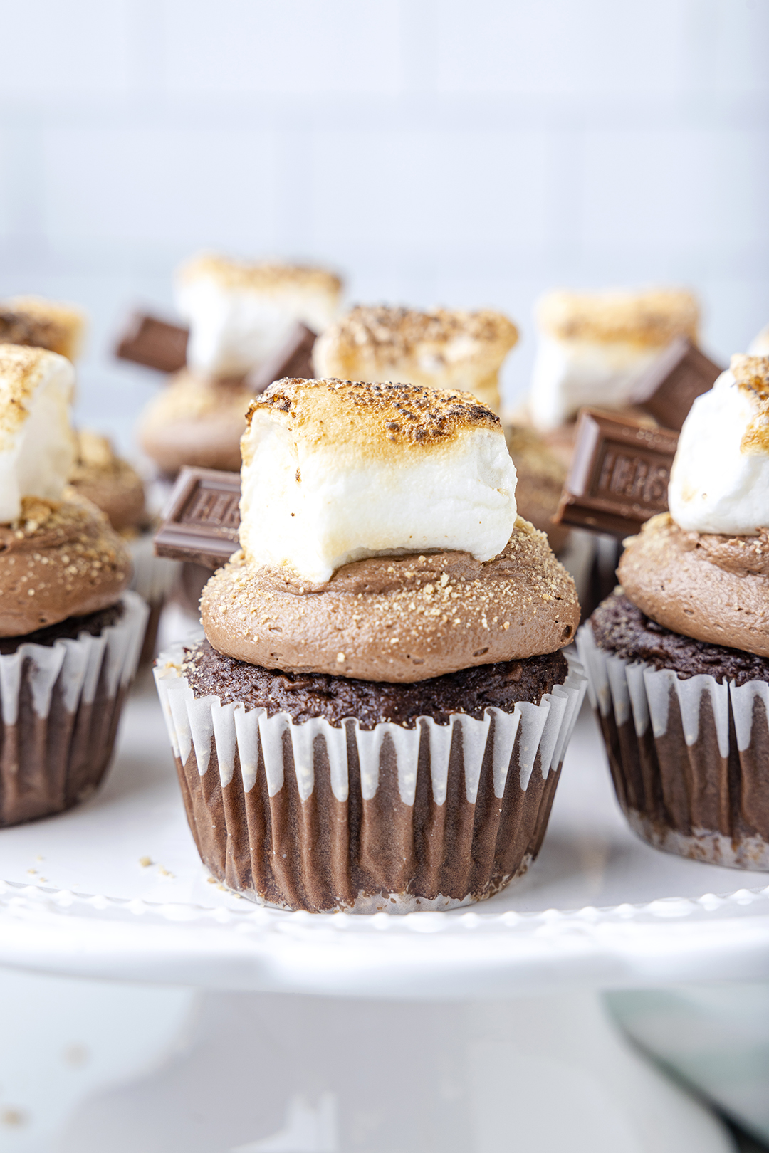 chocolate s'mores cupcakes on a white cake stand. cupcakes are topped with chocolate frosting, toasted marshmallow and mini chocolate bar.