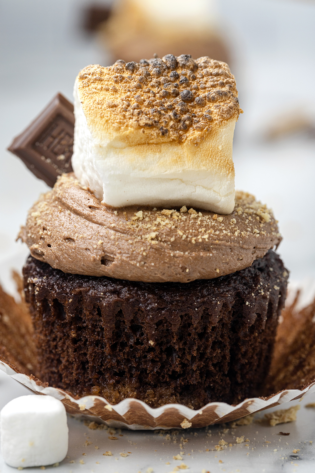 up close view of a s'mores cupcake with graham cracker and chocolate cake base then topped with chocolate frosting, toasted marshmallow, hershey bar and dusted with crushed graham cracker