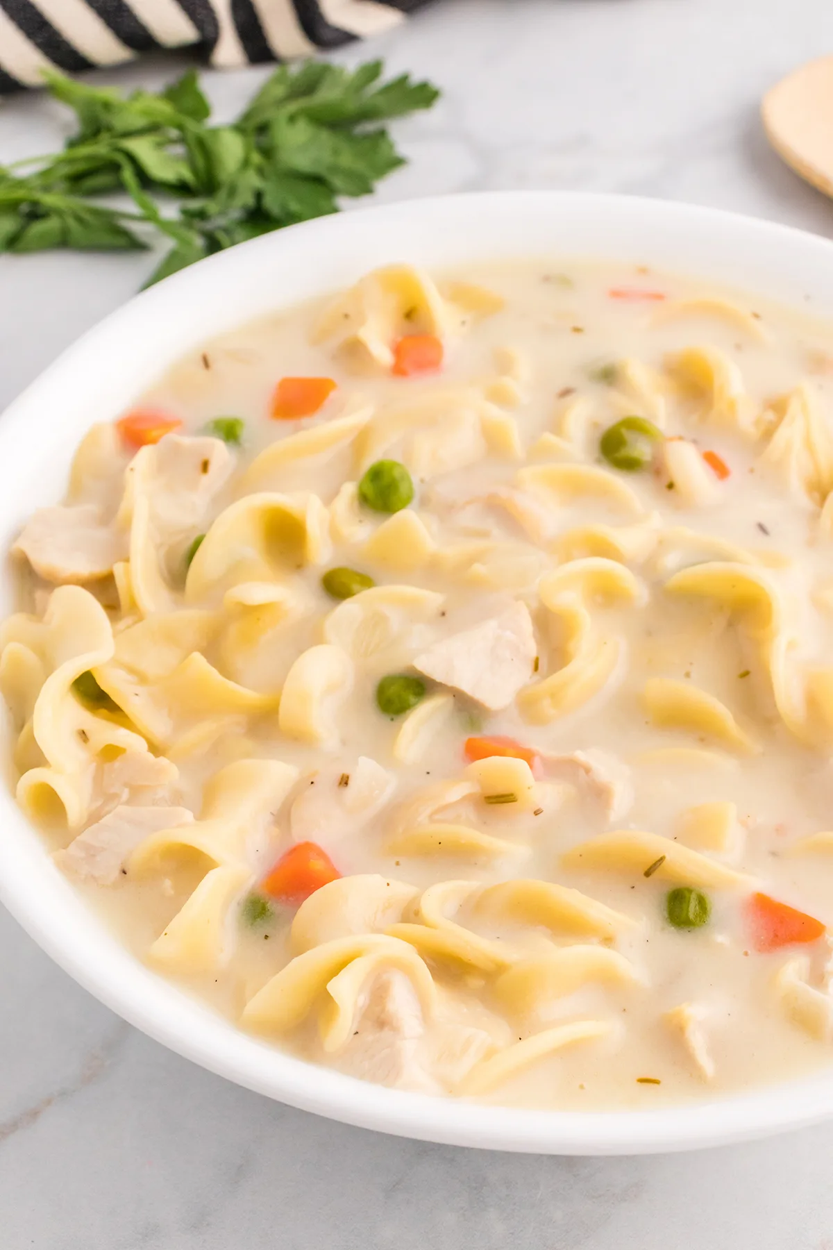 angled down shot of a white bowl filled with creamy chicken noodle soup with peas and carrots
