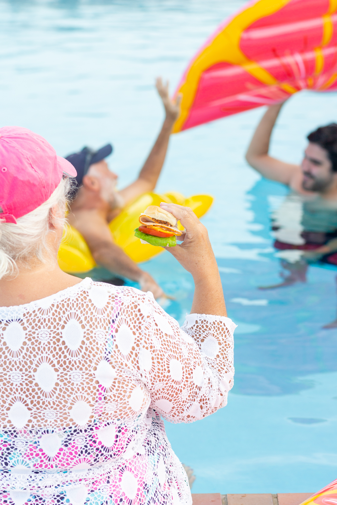 woman enjoying a slider sandwich while sitting with feet in the pool. Family playing with pool floats in the background.