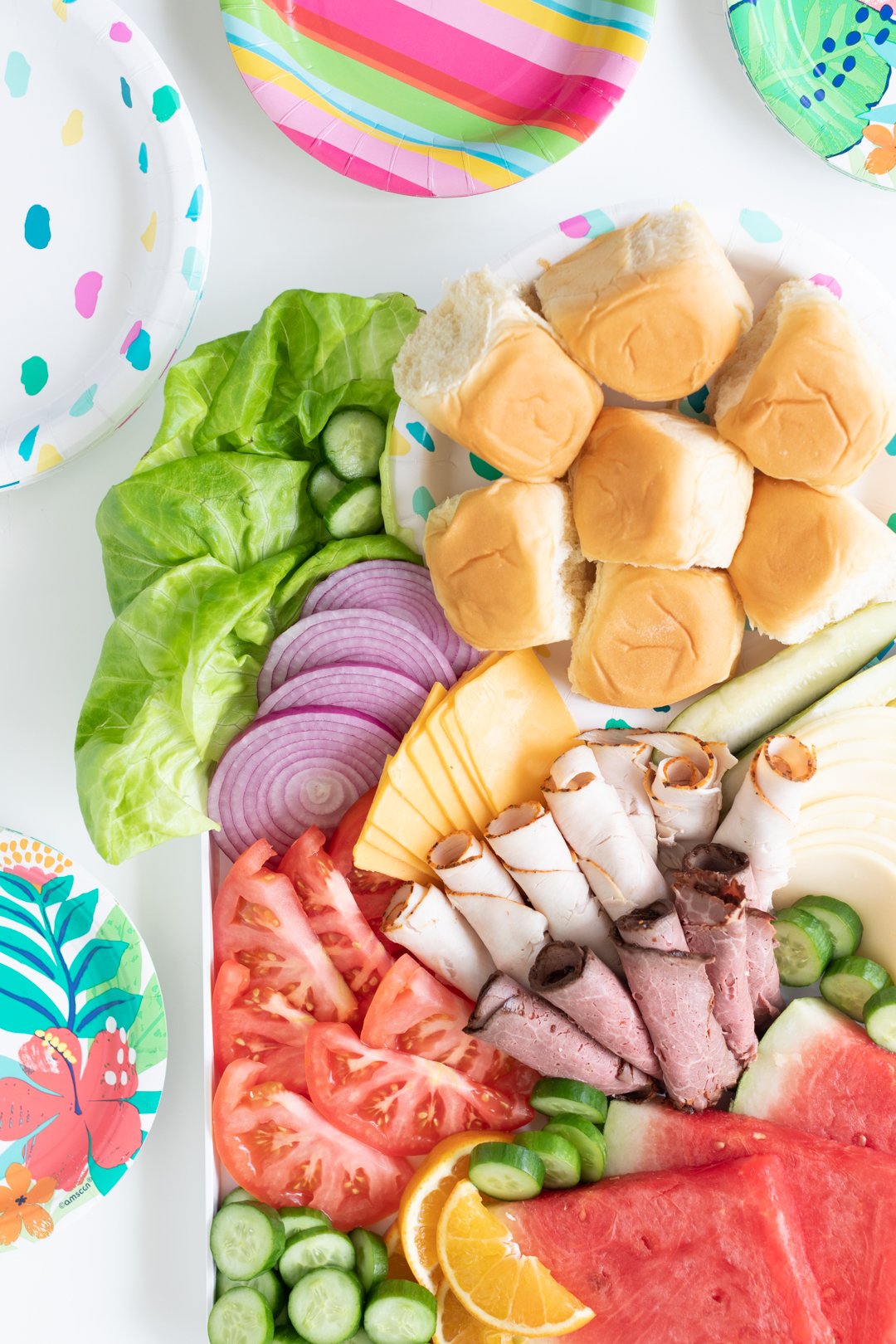 styled sandwich charcuterie board with lettuce, tomato slices, lunch meats, cheeses and fruits with a variety of colorful paper plates surrounding it.