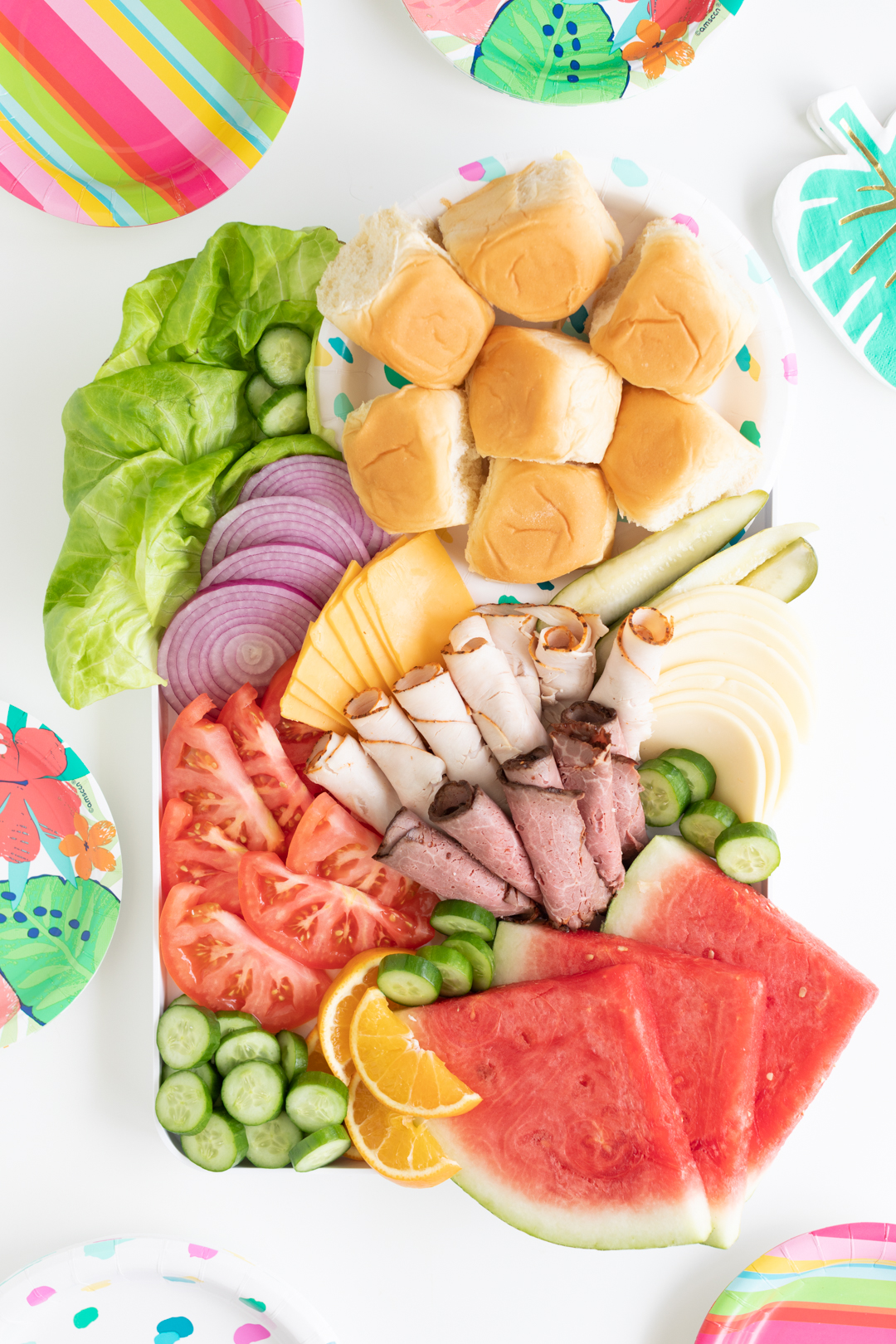 styled sandwich charcuterie board with lettuce, tomato slices, lunch meats, cheeses and fruits with a variety of colorful paper plates surrounding it.