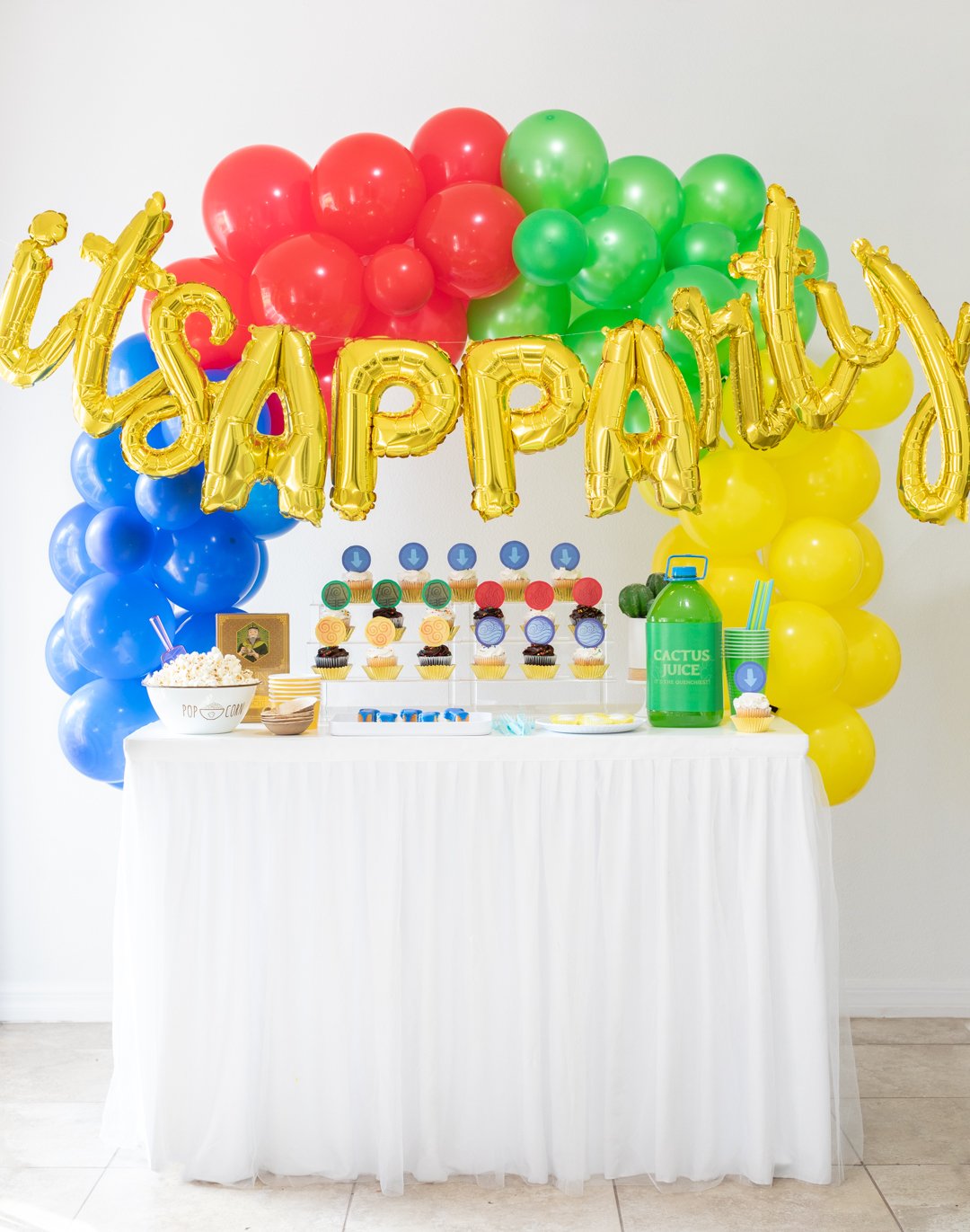 é um APPArty Avatar: The Last Airbender Party Table com balões que dizem "É APPArty"arcos de balões azuis, vermelhos, verdes e amarelos e comida de festa temática.