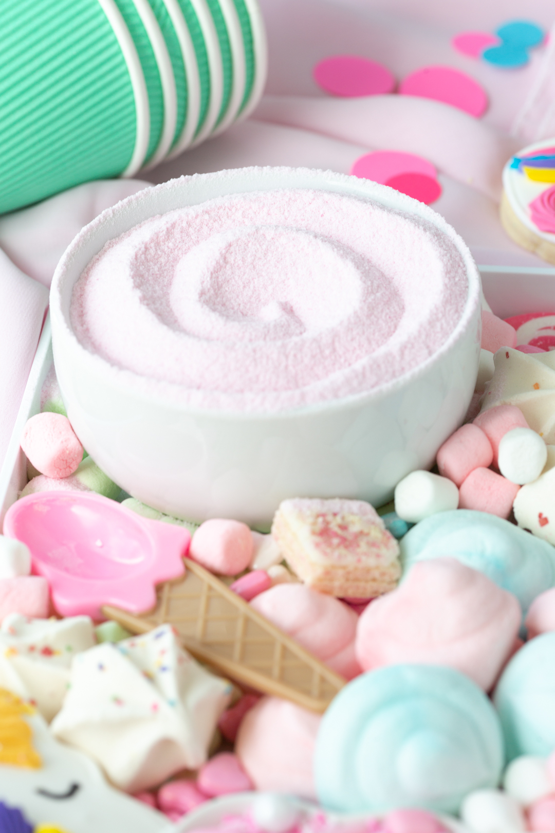 close up view of bowl of pink hot cocoa along with ice cream themed spoon and a variety of marshmallows and small pastel candies