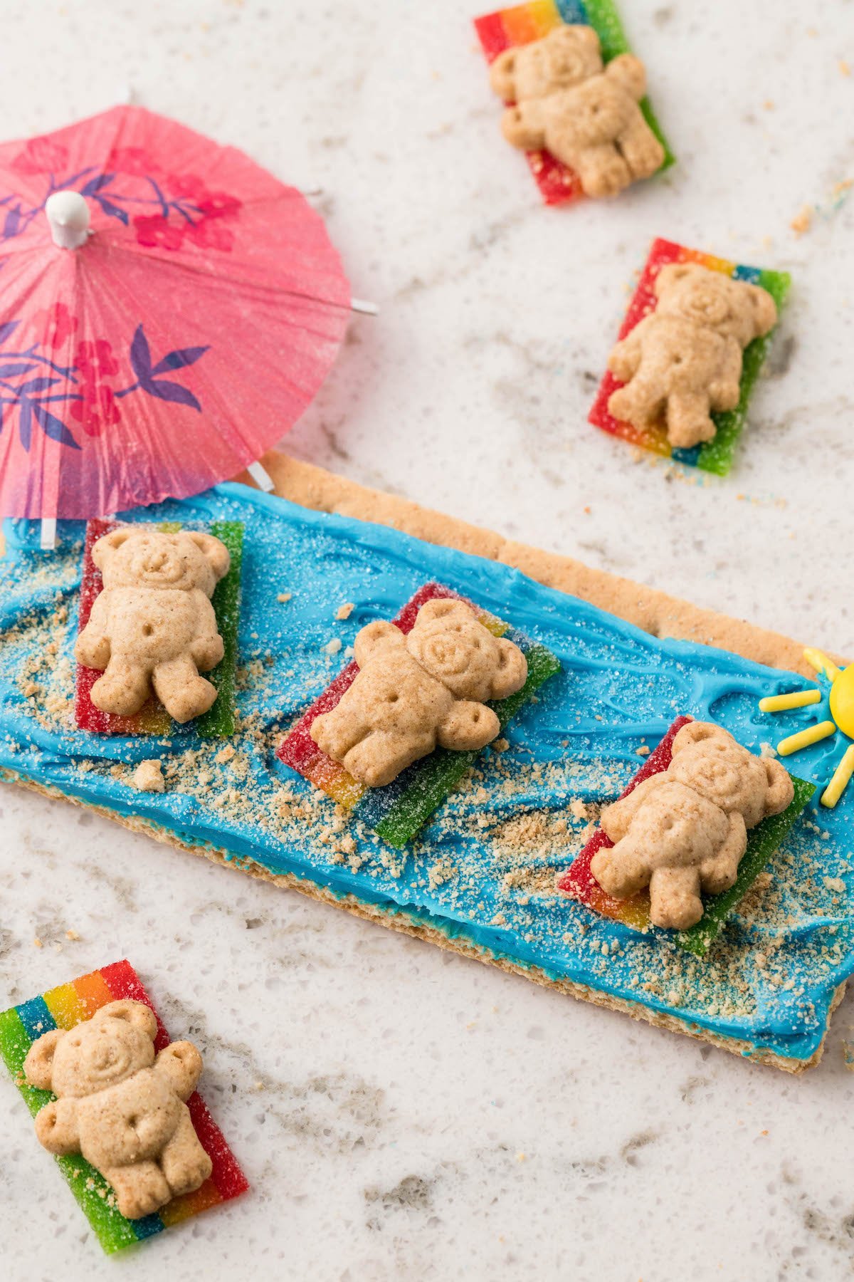 angled down photo of beach day graham crackers with teddy grahams laying on rainbow candy. drink umbrella nearby.