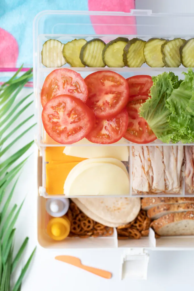 over the top view of a sandwich tackle box with fresh vegetable slices, pickles and lunch meats.